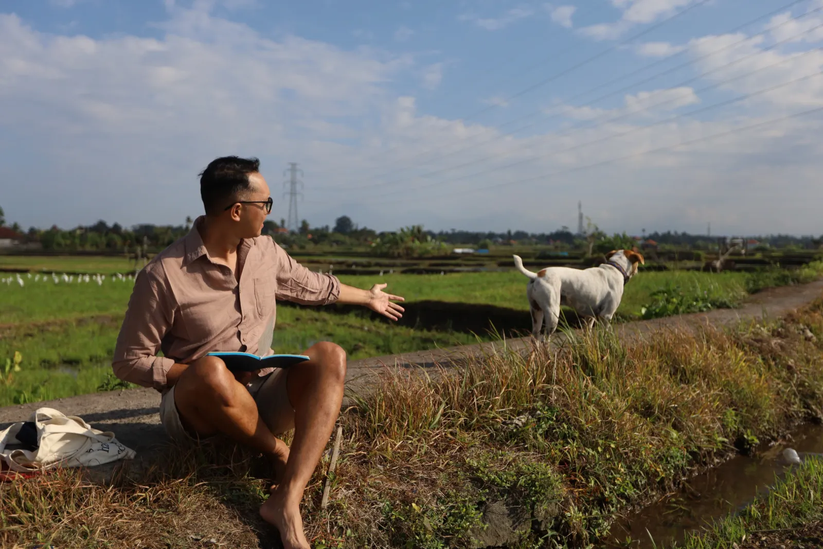 Man in pink shirt with notebook gestures across Bali rice fields, a white dog beside him.